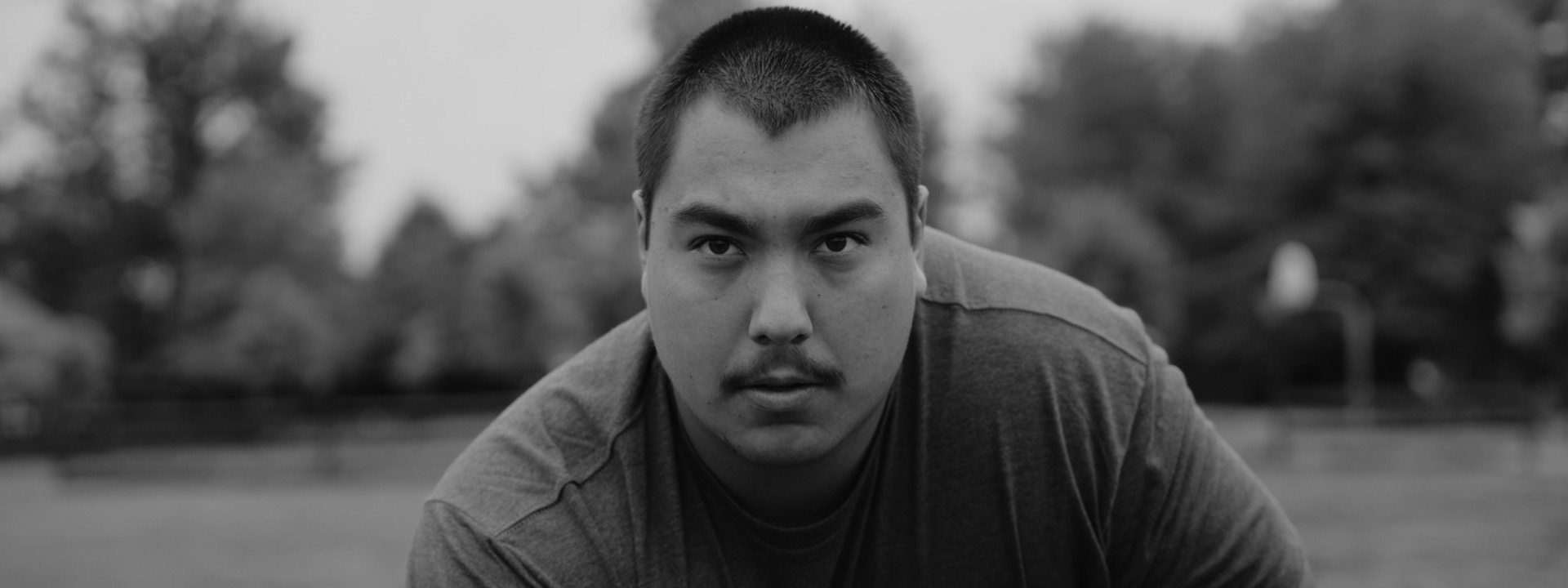 A black and white portrait of a young man leaning toward the camera outdoors, with trees behind him.