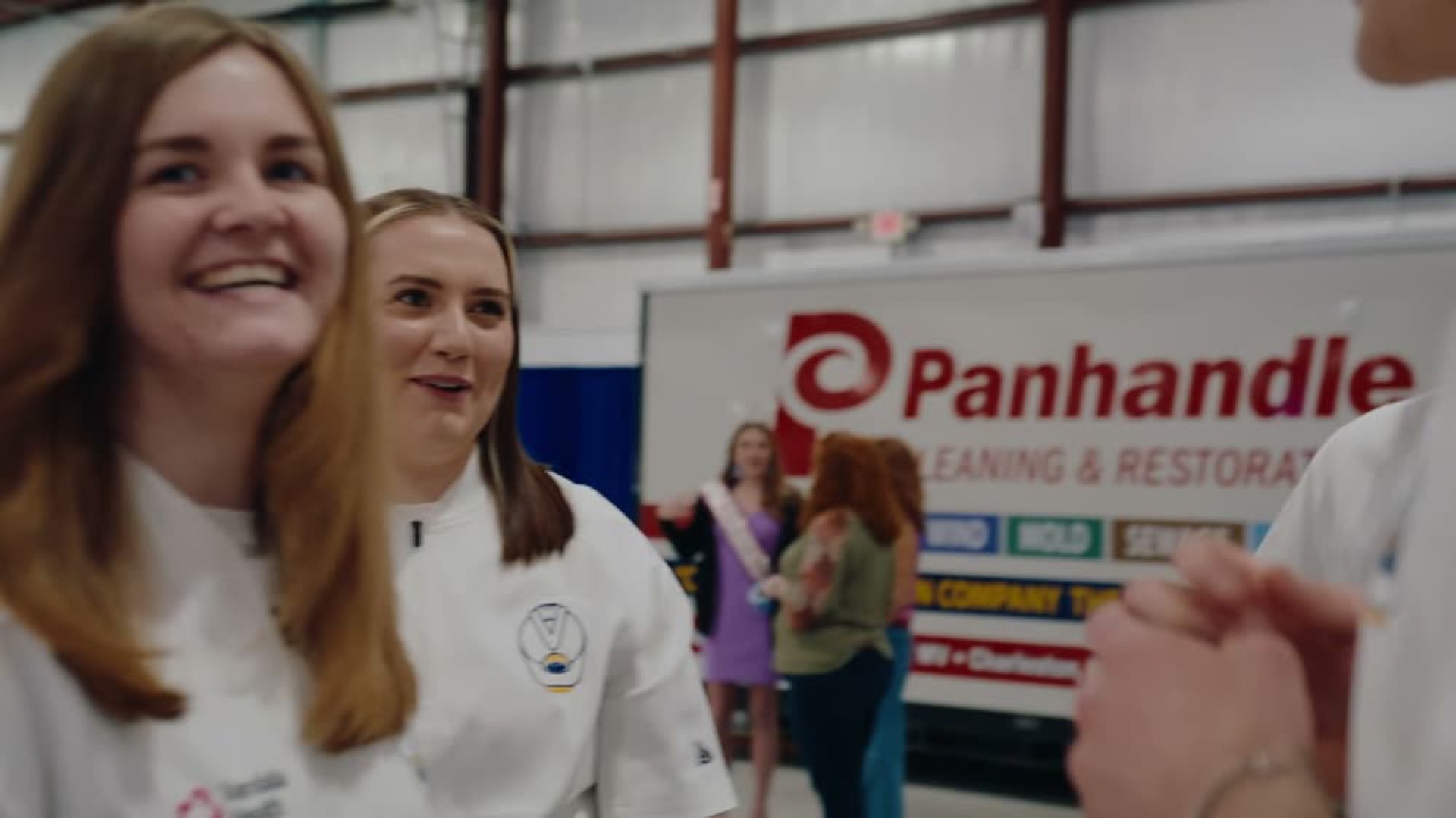 Two smiling young women in white chef coats talk with guests inside a large warehouse hall during an event.
