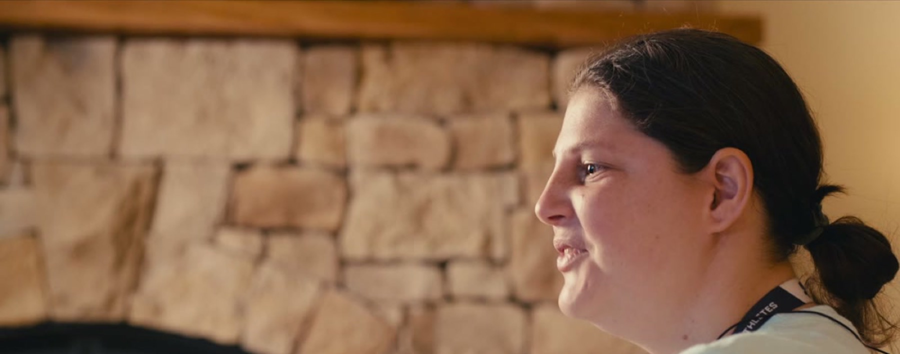 A young woman with her hair pulled back smiles as she talks, seen from the side in front of a stone fireplace.